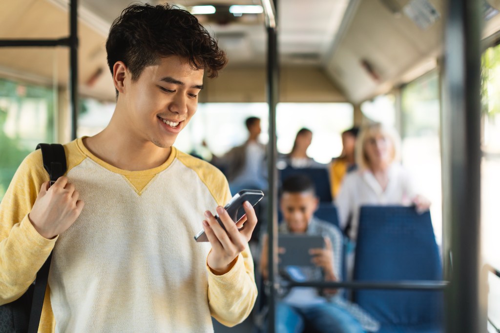 Happy Asian guy using cell reading email messages on display of smartphone connected to public wifi standing in city bus wearing backpack. Male passenger looking at mobile phone screen, texting sms